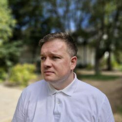 Auto-generated description: A person wearing a white collared shirt stands outdoors with trees and greenery in the background.